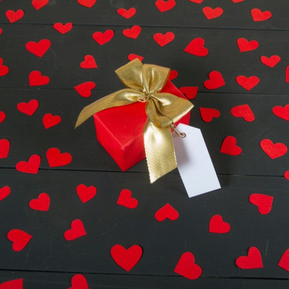 A red box tied with a gold ribbon, sitting on a black background surrounded with red hearts