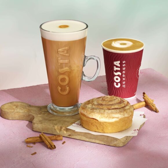 Cinnamon bun lattes from Costa in a glass Costa mug and a takeaway cup behind a cinnamon bun.
