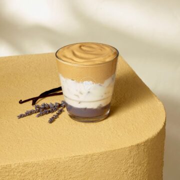 A vanilla lavender iced whipped latte from Costa on a table in front of vanilla beans and sprigs of lavender